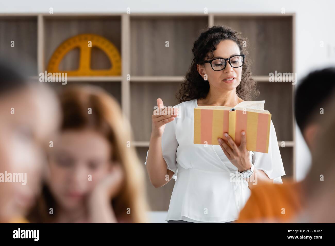 african american teacher reading book near blurred pupils in classroom ...