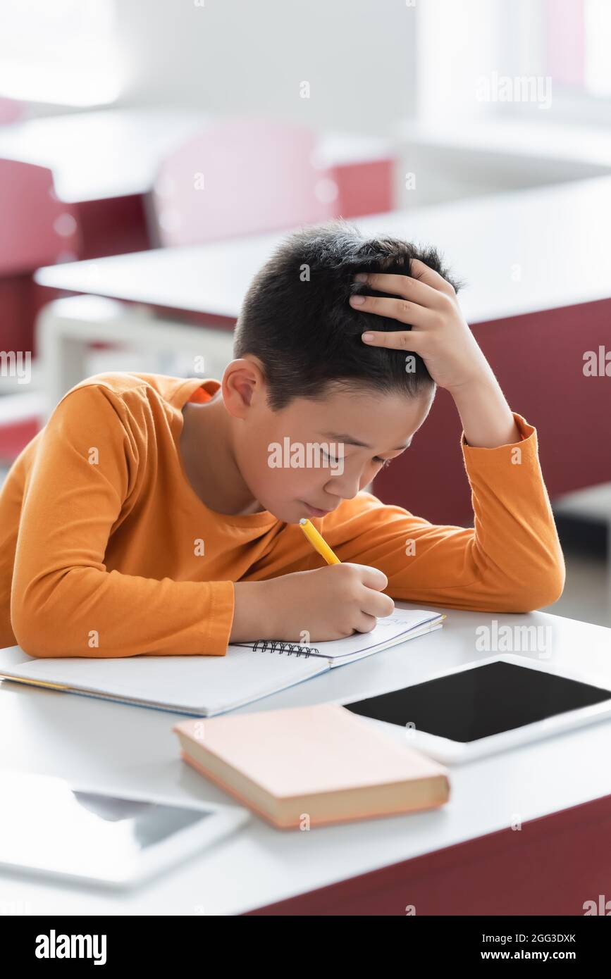 asian boy writing in notebook during lesson near digital tablets on ...