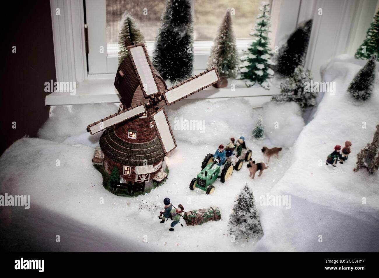 Vintage windmill setting in window in Christmas Village scene with tractor and hayride and kids pulling Yule log and dogs running in the snow - in win Stock Photo