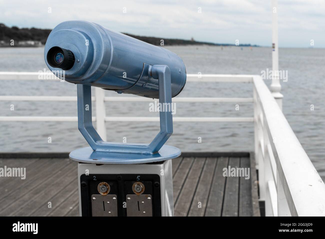Blue telescope mounted on a stand on wooden ocean pier with white railing. Cloudy summer day on the pier of Jurata, Poland. Stock Photo