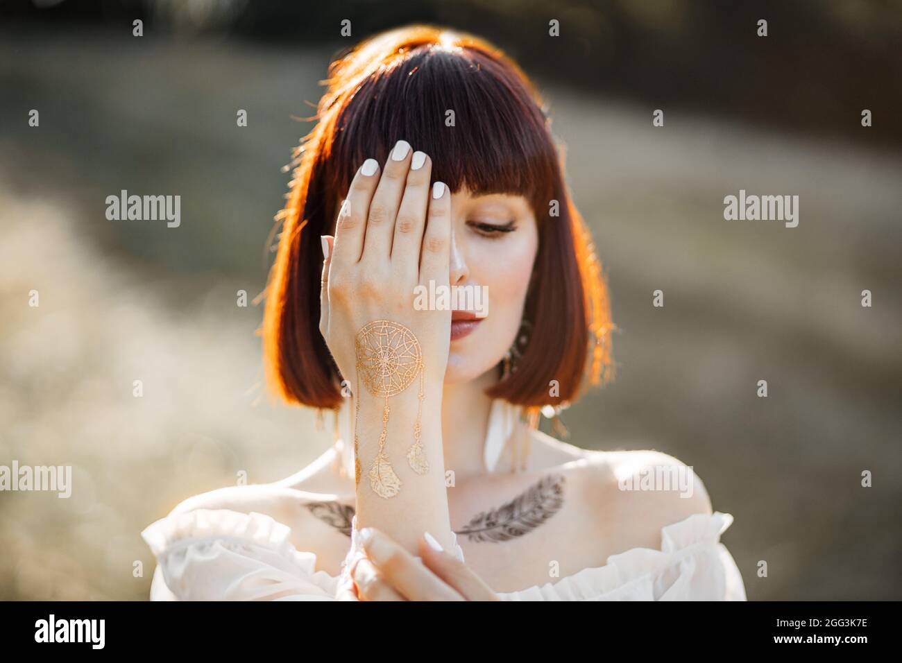 Close up creative portrait of native American Indian woman. Pretty ginger girl with short hair, and feather tattoos on neckline, looking down and covering half of her face with hand Stock Photo