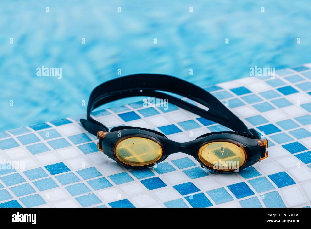 Black professional swimming goggles with water drops on yellow lenses on a blue and white tiles of pool floor. Stock Photo