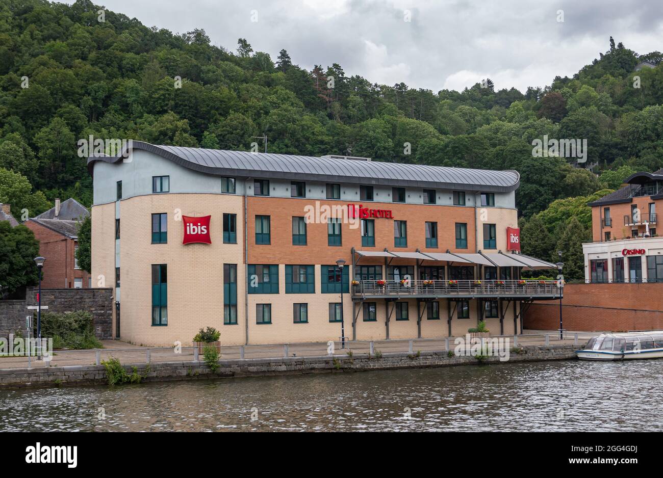 Dinant, Wallonia, Belgium - August 8, 2021: Closeup of IBIS hotel advertises name in red along Meuse river with green forested hill in back under heav Stock Photo