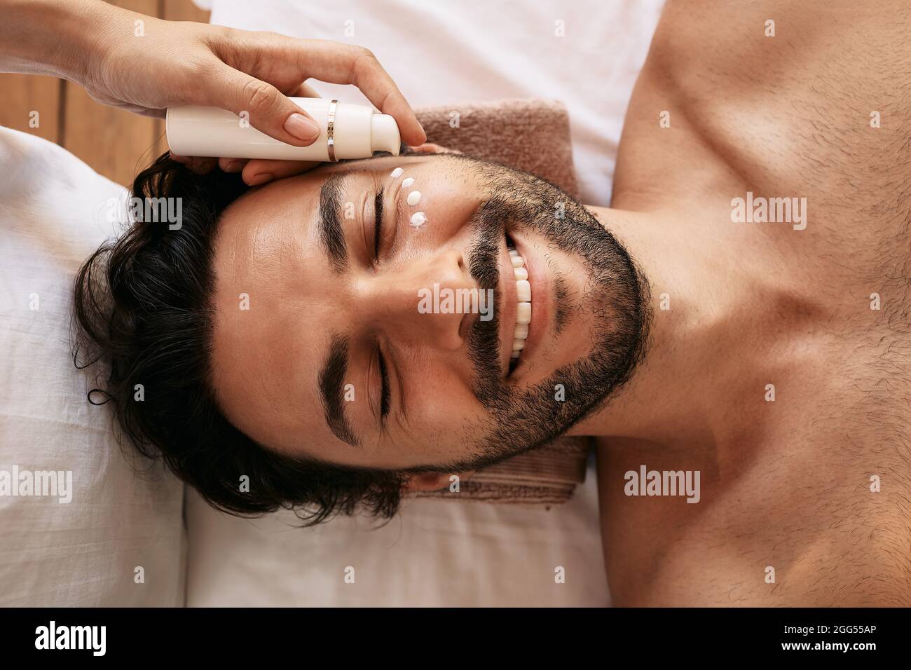 Beautician applies white skin moisturizer to handsome man's face at wellness center. Male patient enjoys beauty treatments, skincare and relaxation at Stock Photo