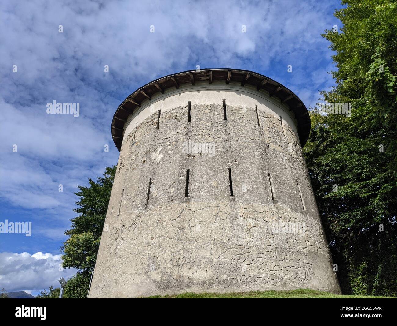 a low angle of white round concrete tower with narrow windows on green lawn, trees under cloudy sky Stock Photo