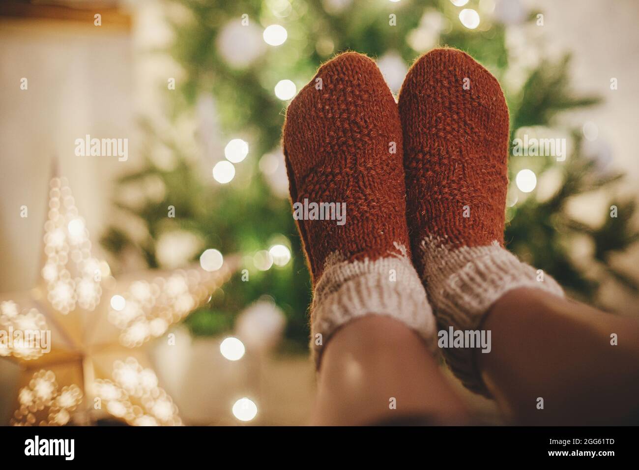 Woman feet in cozy woolen socks on background of christmas tree and star in lights in festive evening room. Cozy winter moments at home. Stylish warm Stock Photo
