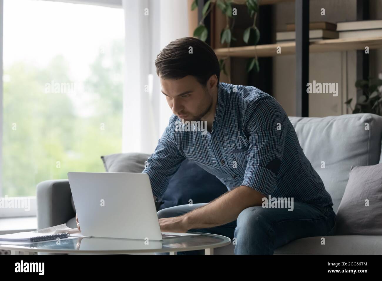 Serious millennial man using laptop, typing, sitting on couch Stock Photo