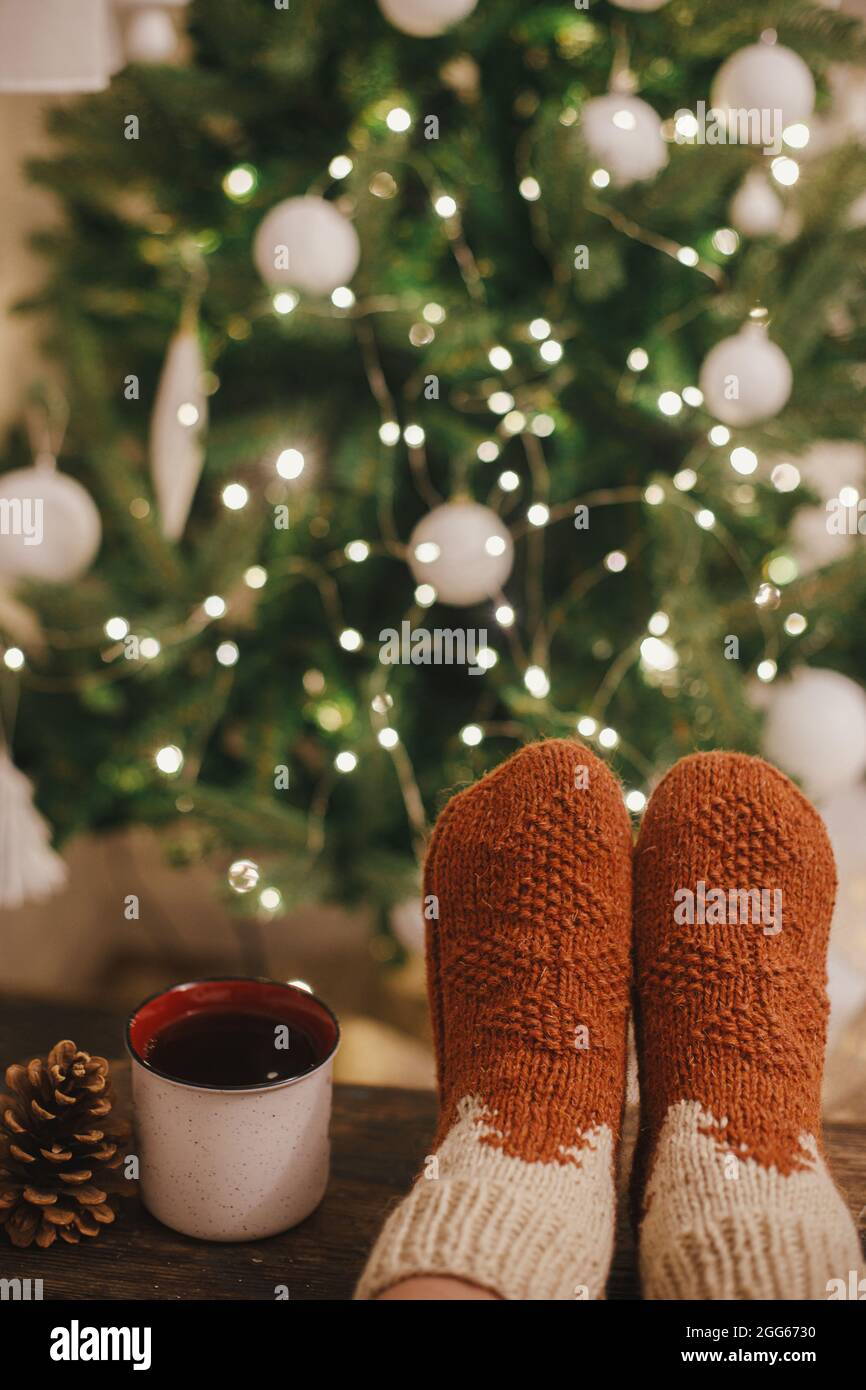 Cozy winter moments at home. Woman feet in cozy woolen socks and cup of warm tea on background of christmas tree in lights in festive evening room. St Stock Photo