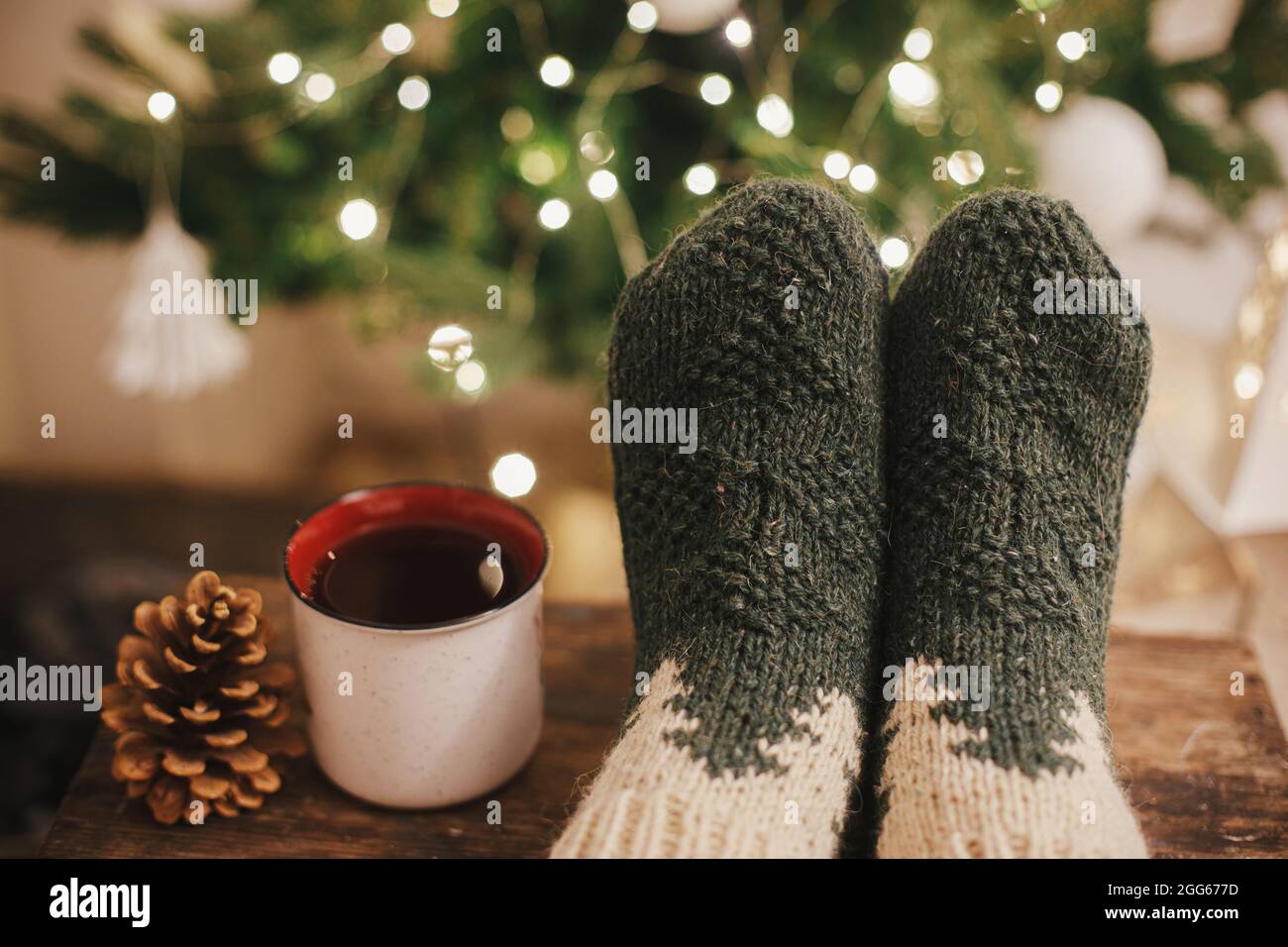 Cozy winter moments at home. Woman feet in cozy woolen socks and cup of warm tea on background of christmas tree in lights in festive evening room. St Stock Photo