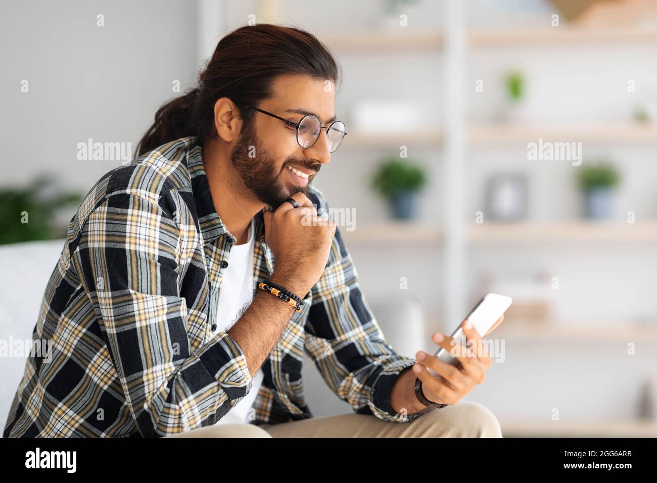 Cheerful long-haired middle-eastern guy hipster sitting on couch, using smartphone Stock Photo
