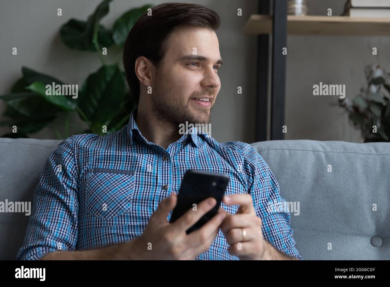 Positive thoughtful cellphone user sitting on sofa at home Stock Photo