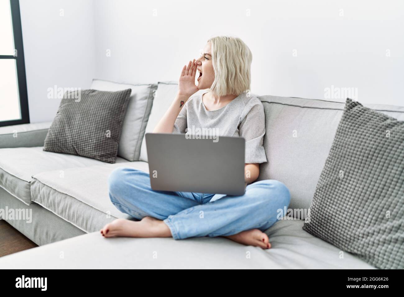 Young caucasian woman using laptop at home sitting on the sofa shouting ...