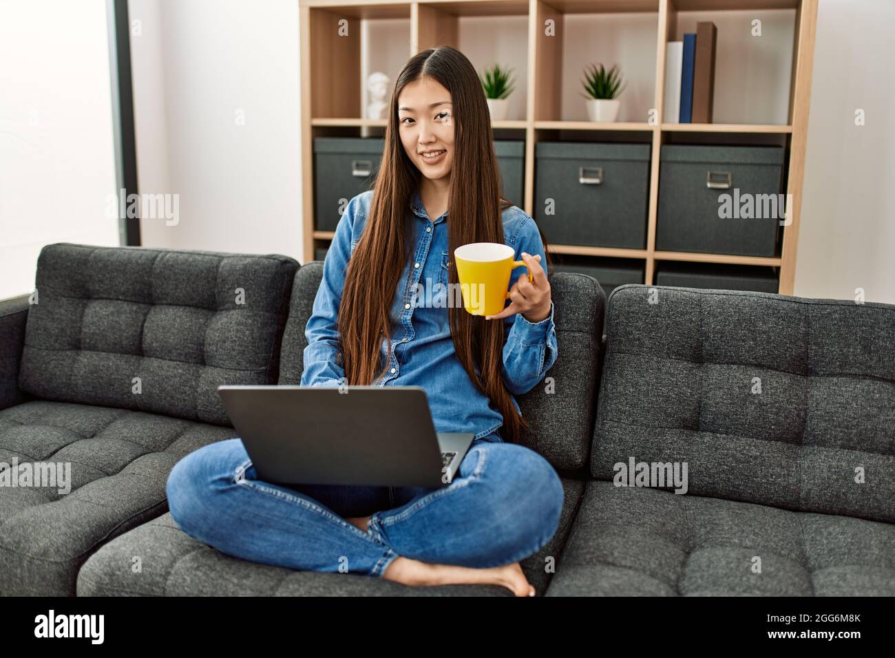 Young chinese girl using laptop and drinking coffee sitting on the sofa ...