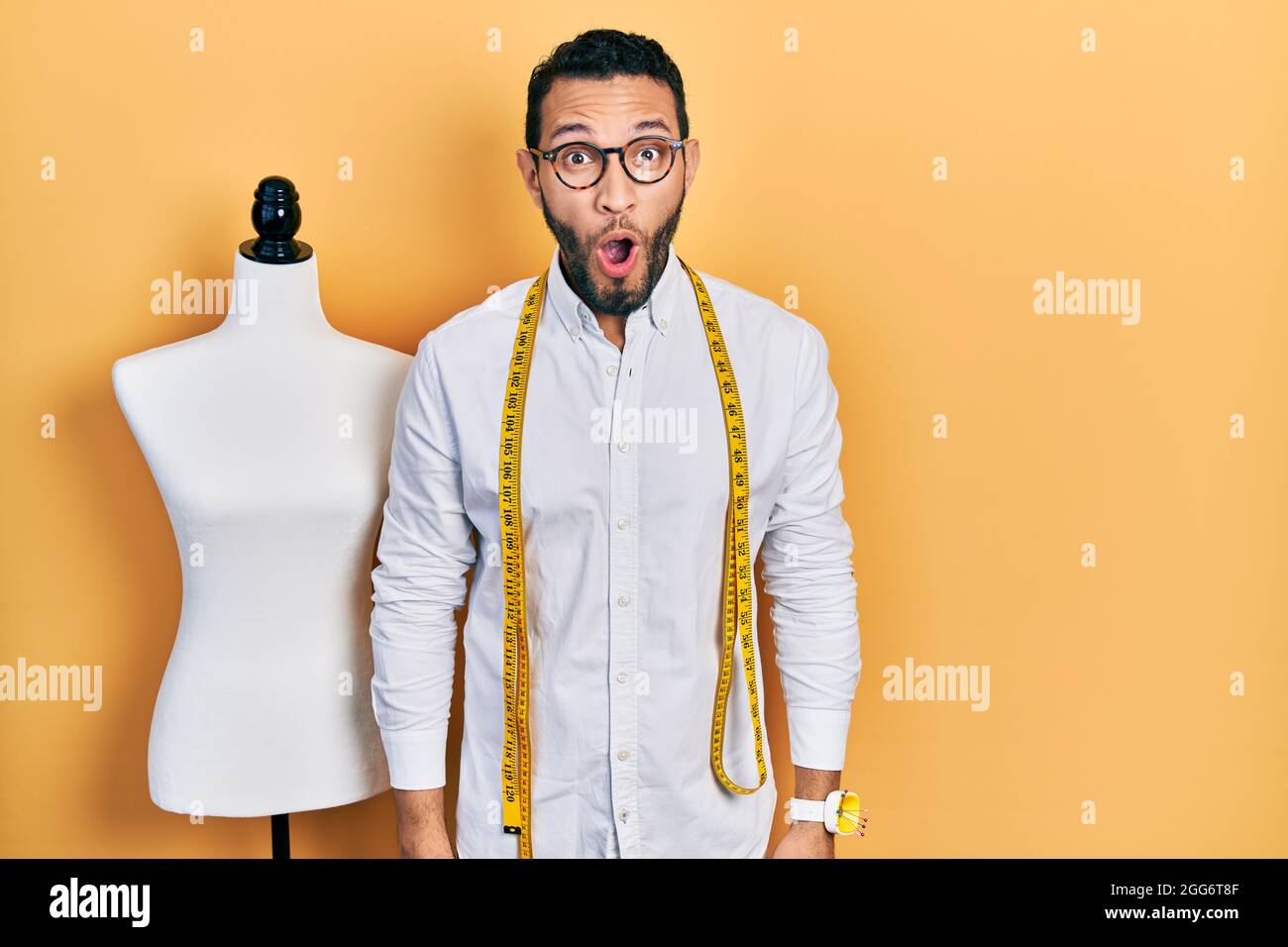 Hispanic man with beard dressmaker designer standing by manikin scared and amazed with open mouth for surprise, disbelief face Stock Photo