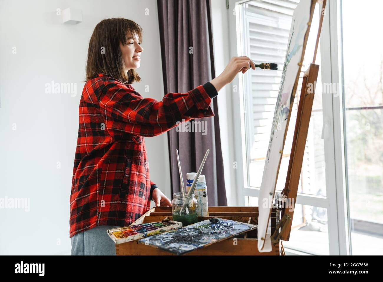 Smiling young white woman in casual wear painting on a canvas indoors ...