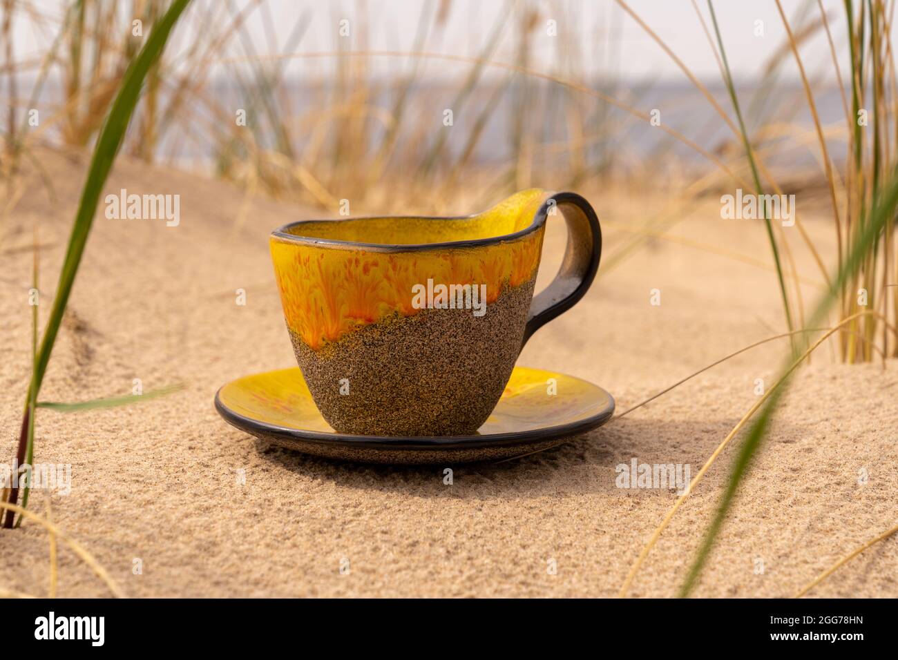 A clay cup with a saucer is placed in the sand of the beach between the bends.  A clay cup with a yellow glass glaze and a matching saucer is placed i Stock Photo