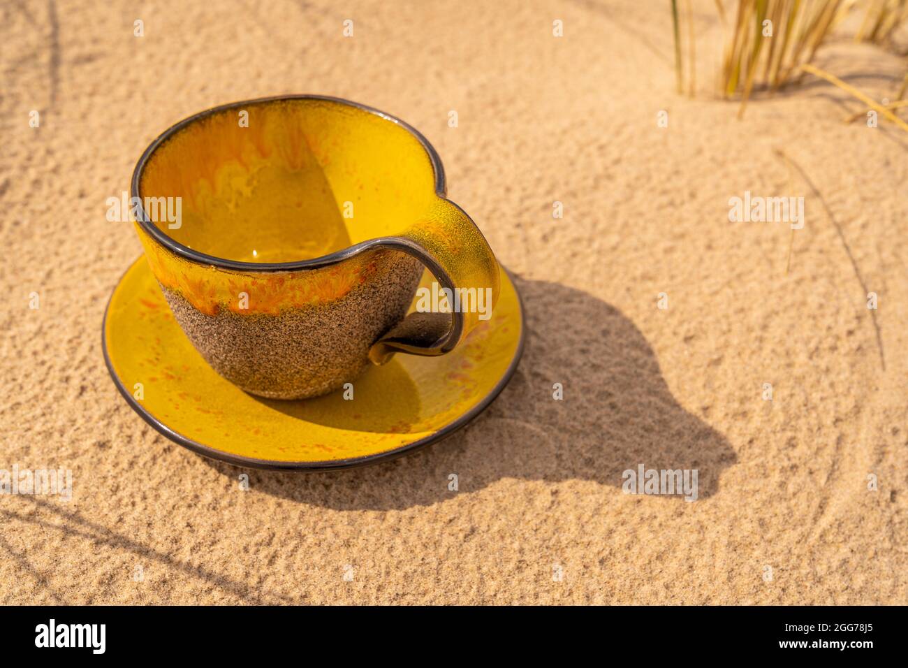 A clay cup with yellow glass glaze and a matching saucer is placed in the beach sand.   Oblique views of the cup with a saucer. Stock Photo
