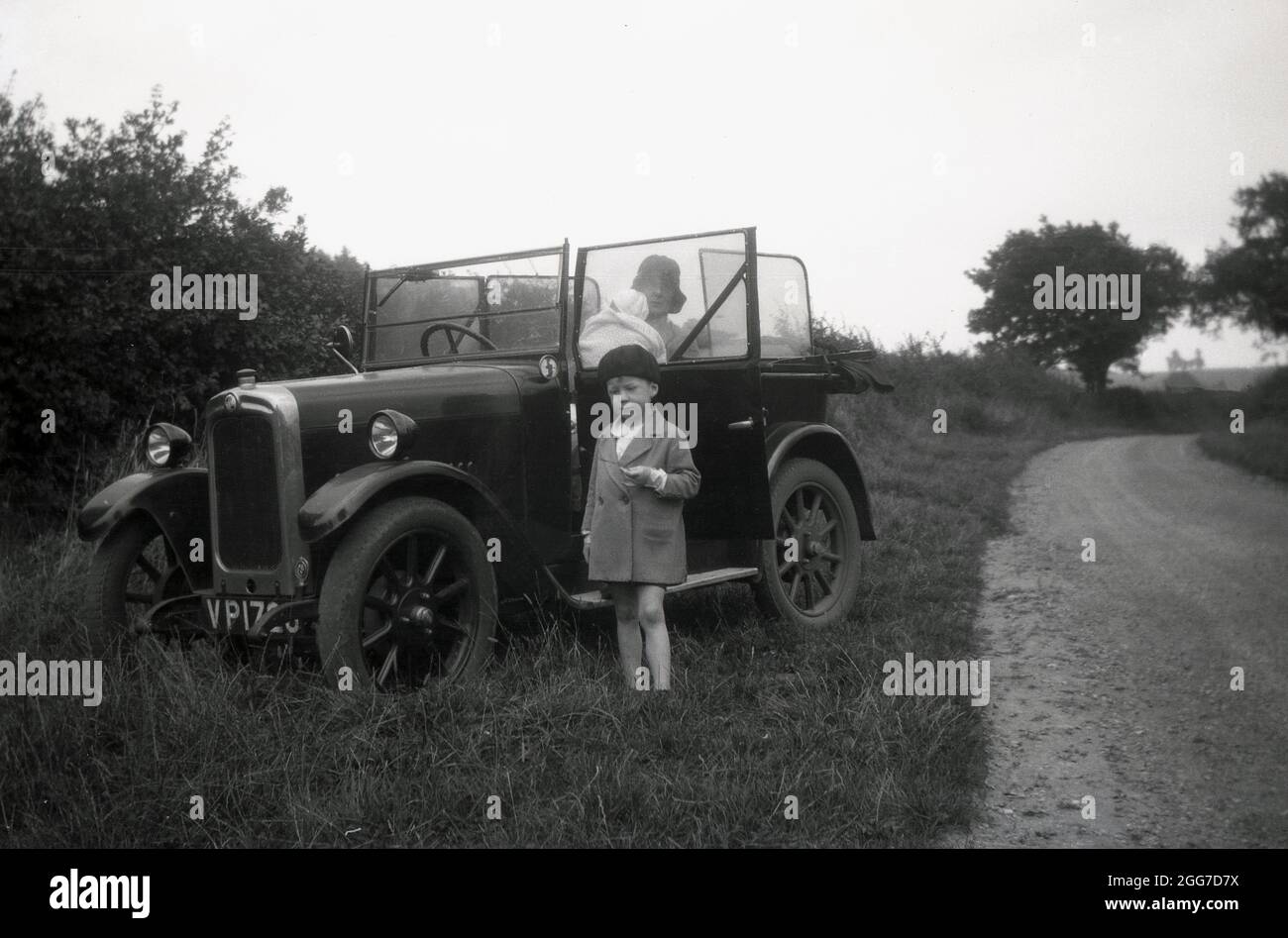 circa 1930s, historical, on a grass verge beside a country lane, a lady sitting in the passenger seat of an open-top car of the era, holding her baby, with her young son is standing infront of the car. Stock Photo