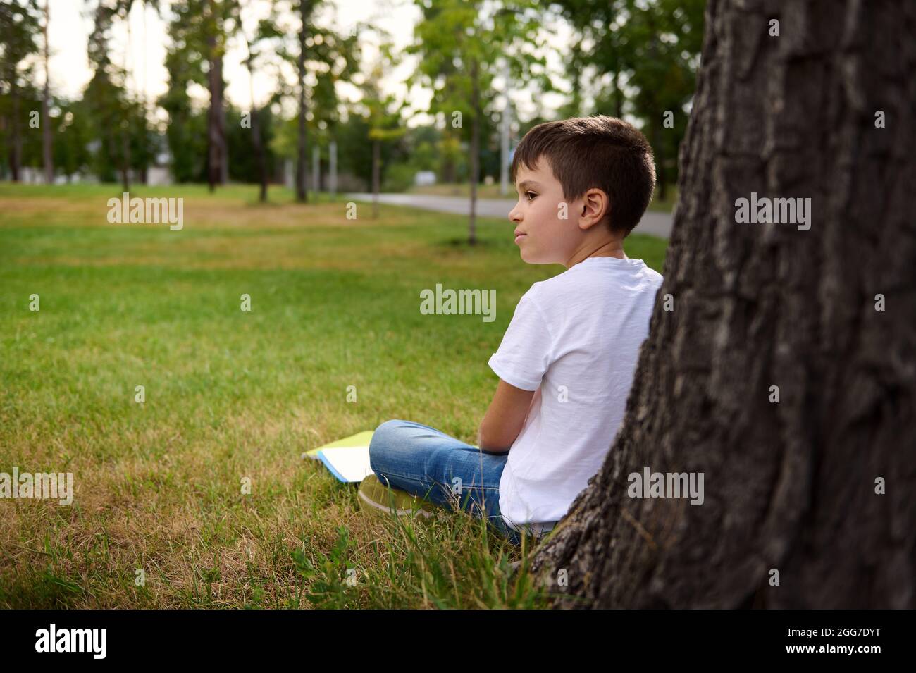 Side view of distracted schoolboy enjoying recreation after school , sitting on the grass of the park, leaning against a tree, doing his homework and Stock Photo