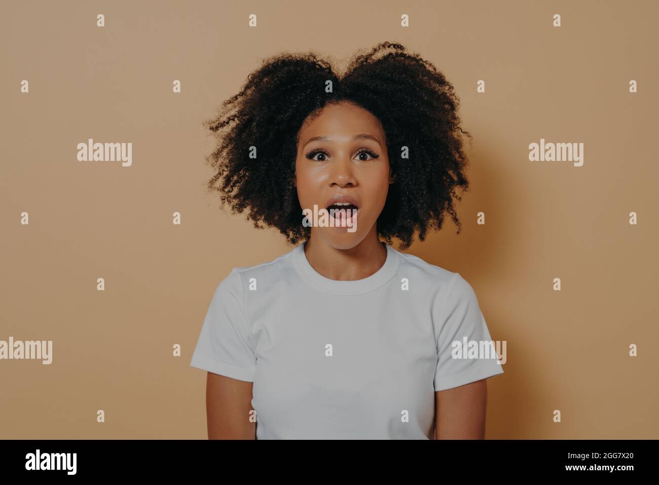Amazed stunned dark skinned female looking at camera with opened mouth isolated over beige wall Stock Photo