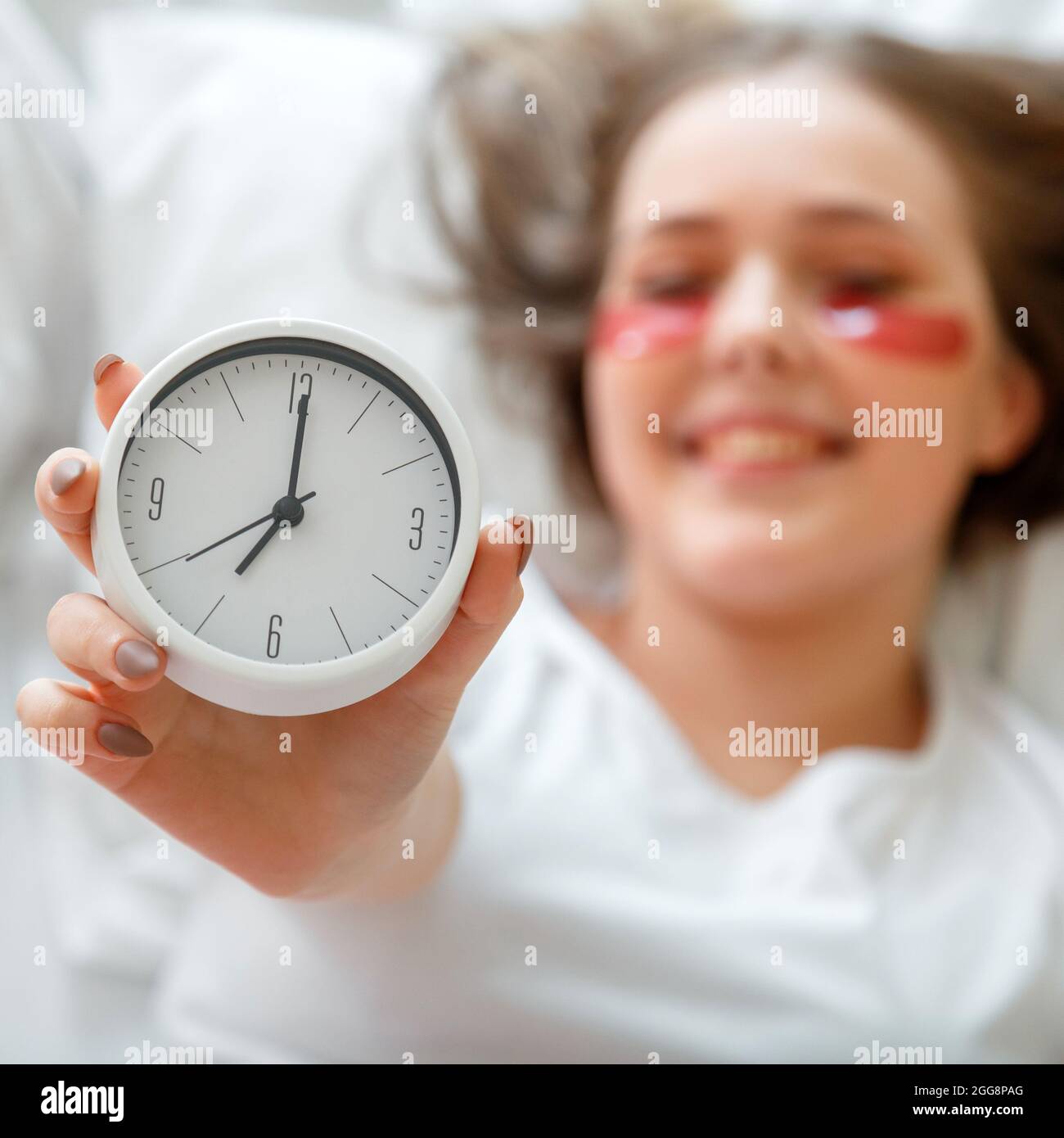 Alarm clock in female hand. Happy young woman wake up after sleeping in bed holding side alarm clock. Morning beauty routine. Woman use eye patches Stock Photo