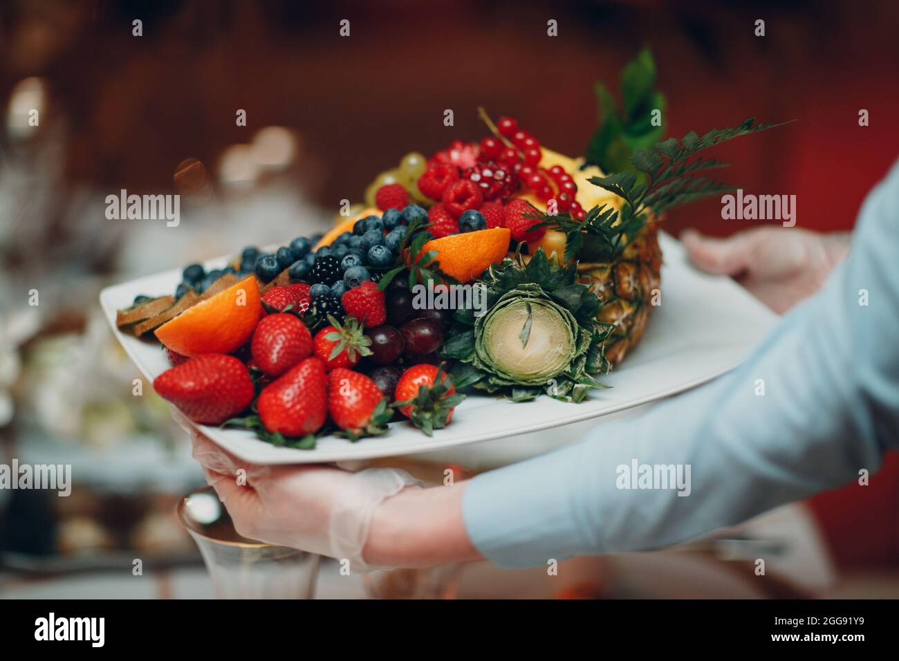 a waiter in a restaurant holds a white plate with different fruits pineapple strawberries berries Healthy food Concept. Stock Photo