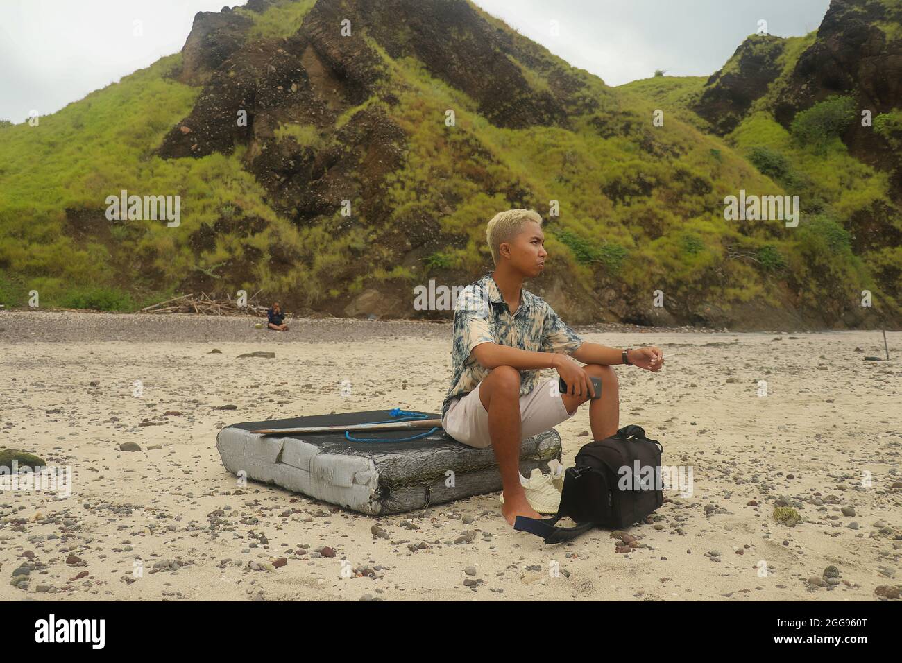 A young man sits on a board with hills on the background in Padar Island. Stock Photo