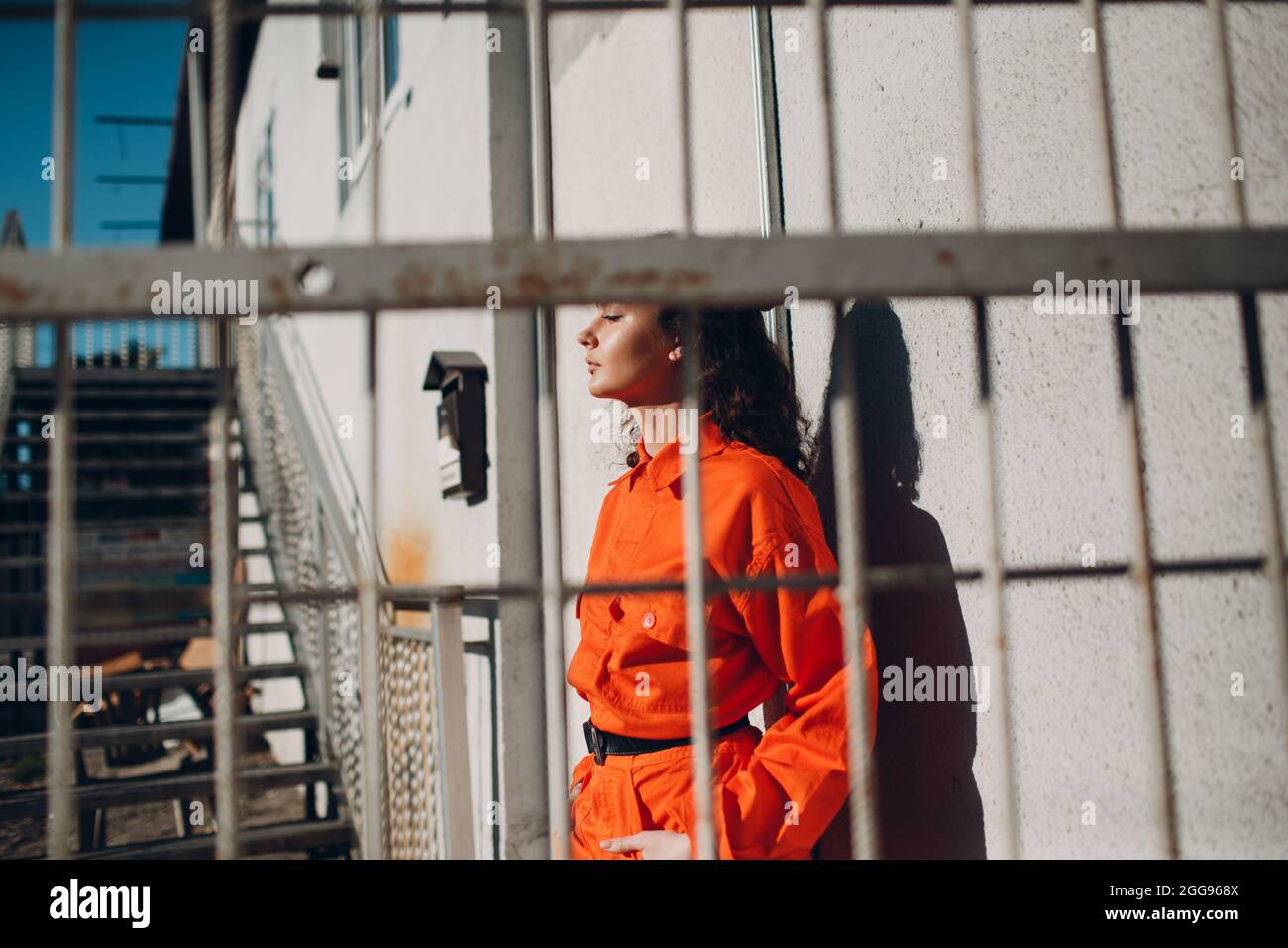 Young brunette curly woman in orange suit behind cage. Female in colorful overalls portrait Stock Photo