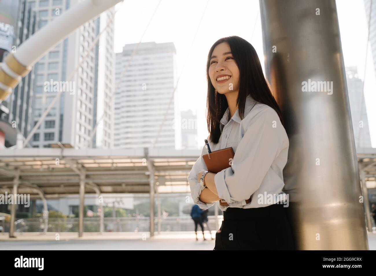Cheerful beautiful young Asian woman standing leaning against building pillar holding a notebook and pen while looking away Stock Photo
