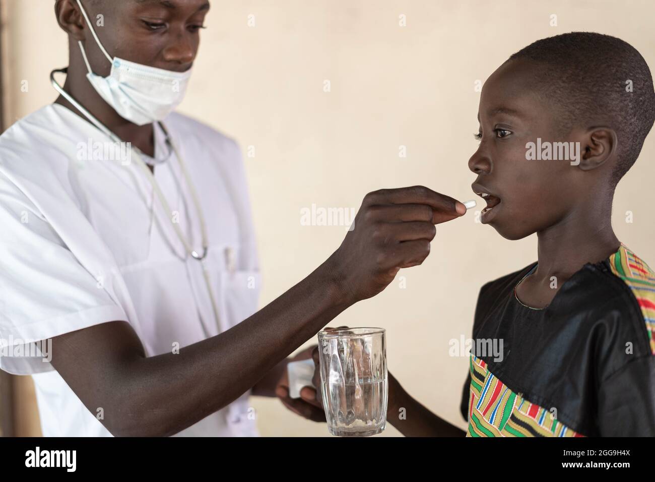 Health professional with protective mask and stethoscope giving a pill to a smal black boy holding a glass of water; child health care concept Stock Photo