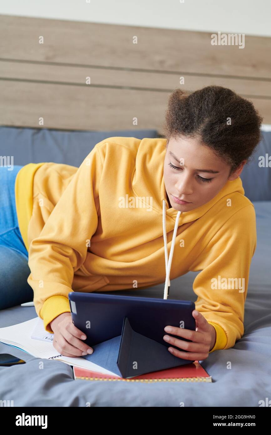 Serious teenage girl in yellow hoodie lying on bed in her room and