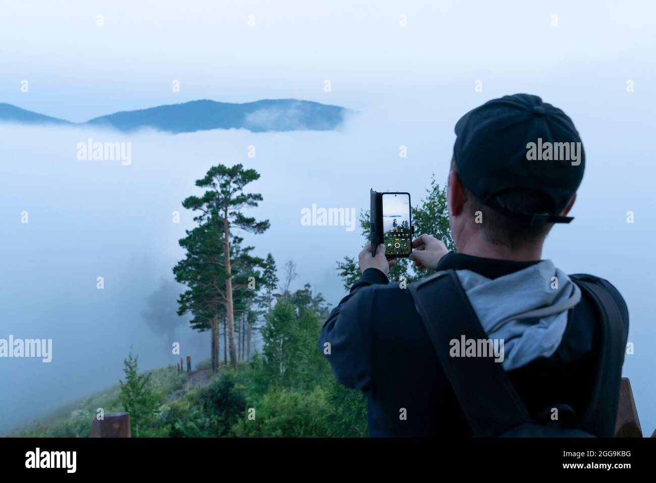 a male tourist in a cap takes photos of the morning uman and his journey on his mobile phone. early morning in the gloomy dramatic forest. Stock Photo