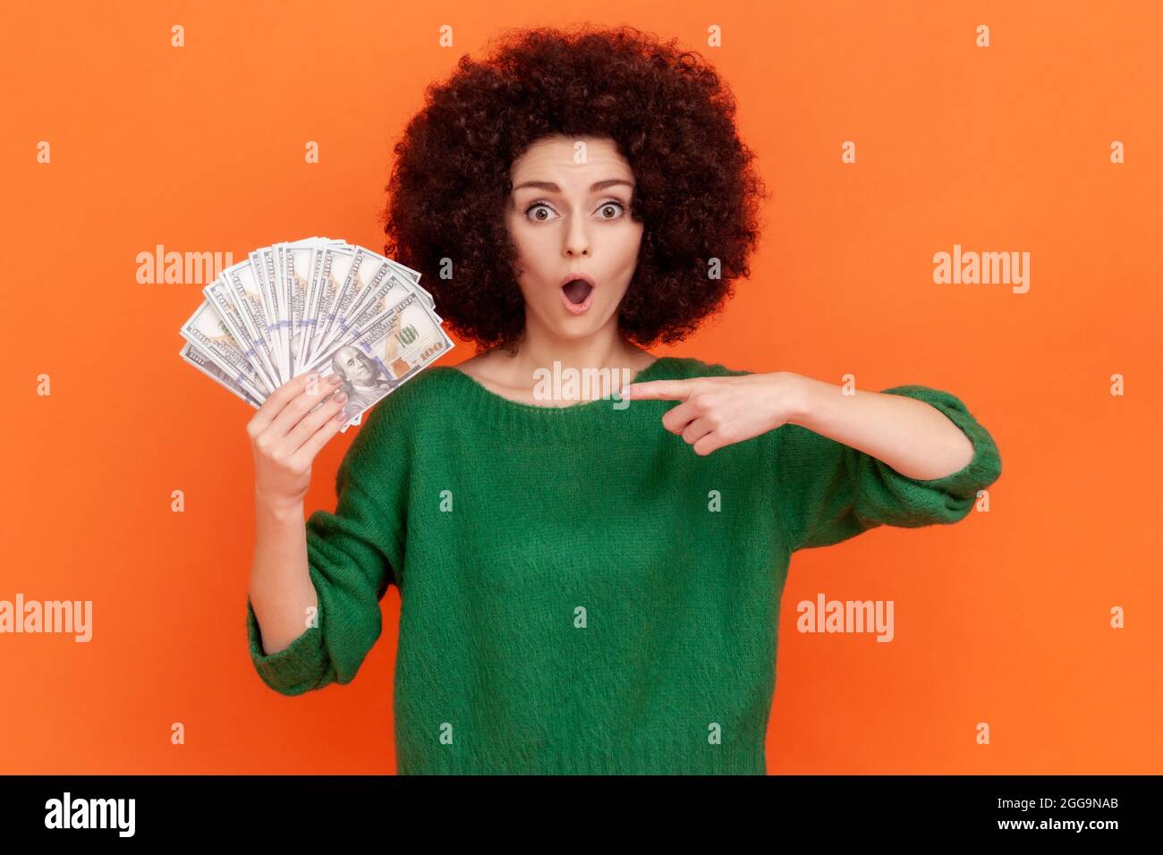 Portrait of shocked woman with curly hair wearing green casual style sweater holding fan of dollars and pointing at banknotes with open mouth. Indoor Stock Photo