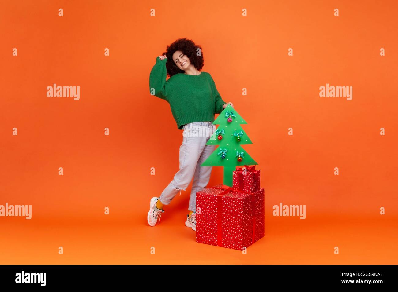 Full length portrait of woman with curly hair in green casual style sweater standing with present boxes and decorative Christmas tree, celebrating. In Stock Photo