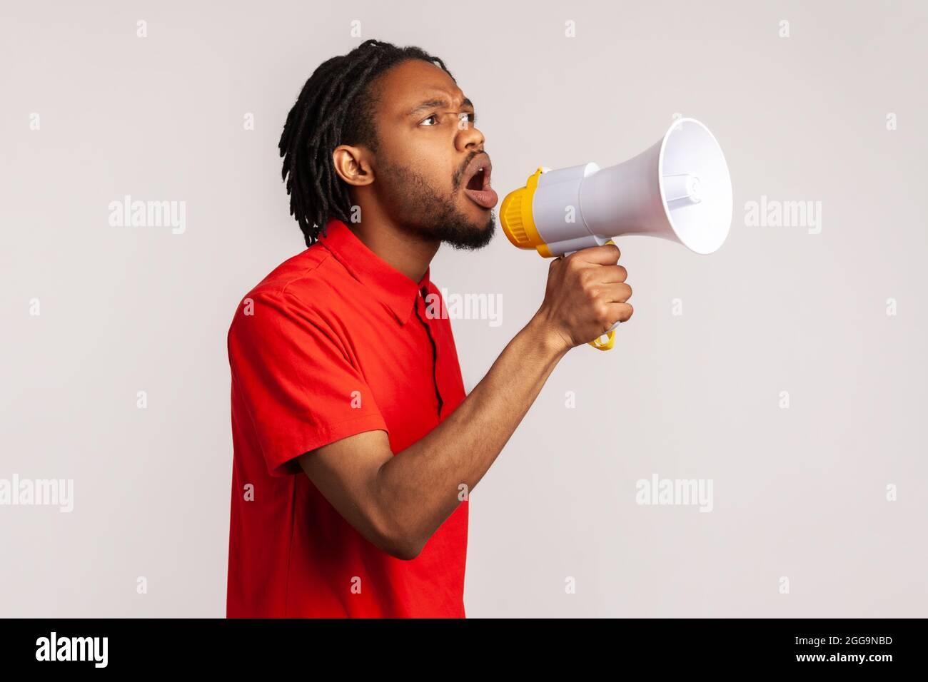 Side view of man with dreadlocks wearing red casual style T-shirt, speaking holding loudspeaker near mouth, talking about new important information. I Stock Photo