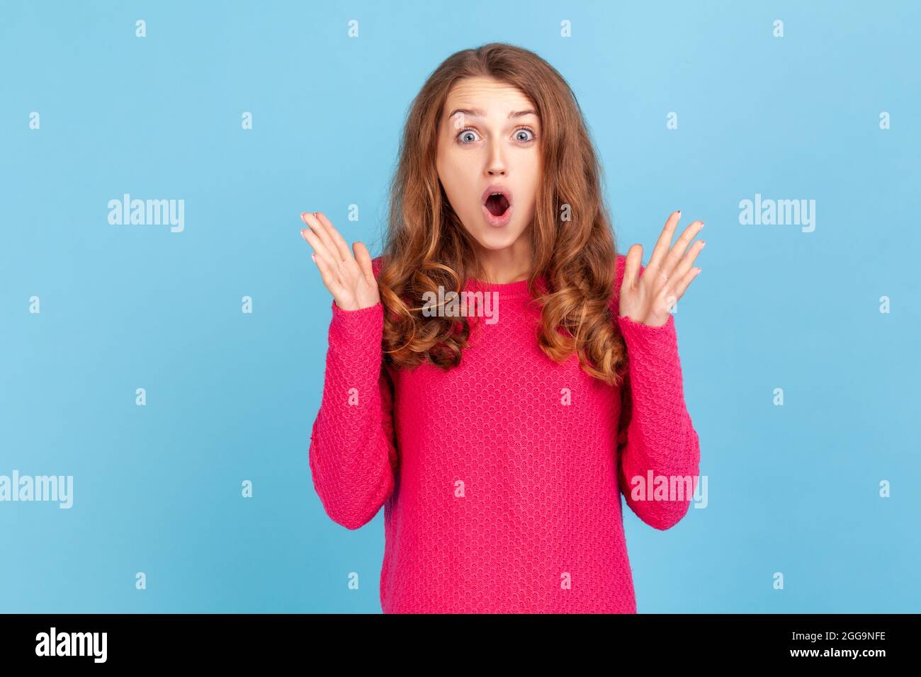 Portrait of shocked woman with wavy hair wearing pink pullover standing with raised arms and open mouth, hearing astonishing news, saying wow. Indoor Stock Photo