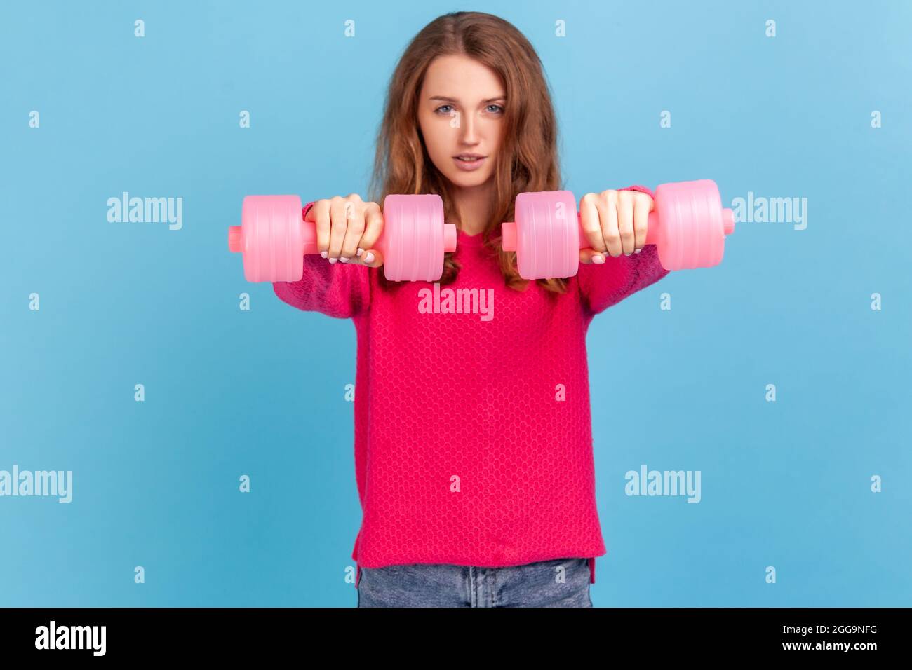Winsome confident woman with wavy hair wearing pink pullover standing holding out two dumbbells, looking at camera, training her biceps. Indoor studio Stock Photo