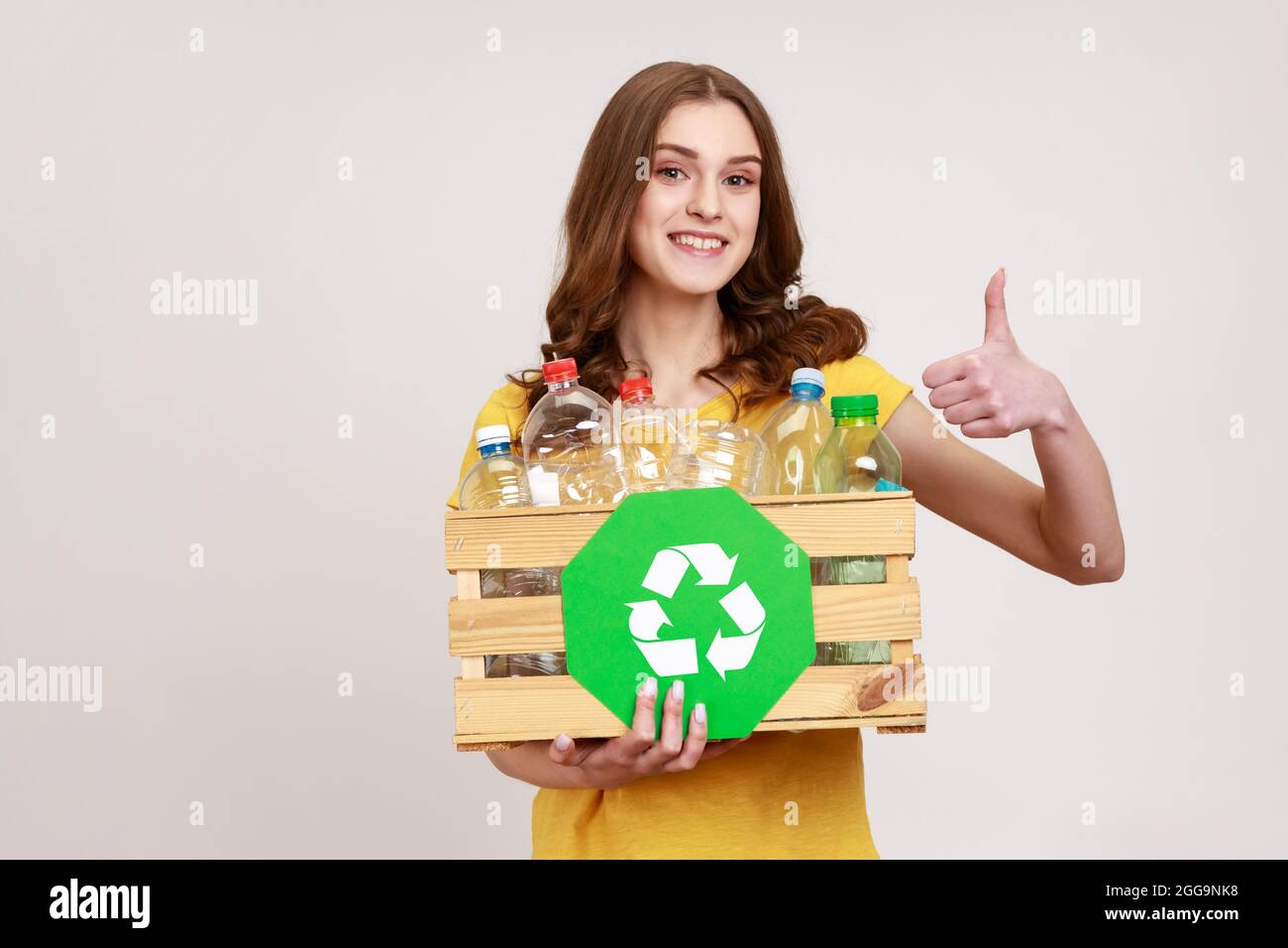 Recycling, waste sorting and sustainability concept. Smiling young female in yellow t-shirt holding box with plastic bottles and showing thumb up. Ind Stock Photo