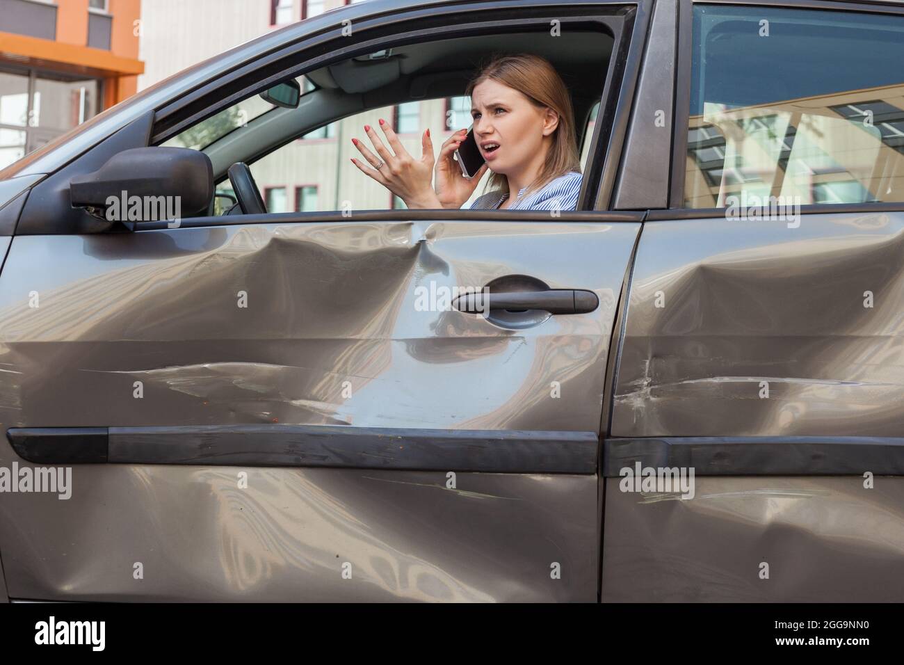 Profile portrait of blonde woman with frowning face sitting behind wheel of damaged car and talking phone, raising palms, explaining detail of road ac Stock Photo