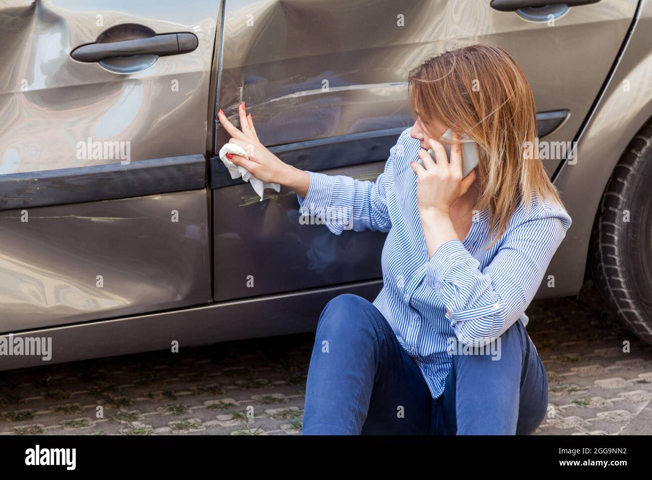 Blonde female in casual attire sitting near dented car and touching scratches and dents on automobile door, talking phone with insurance agent or ask Stock Photo