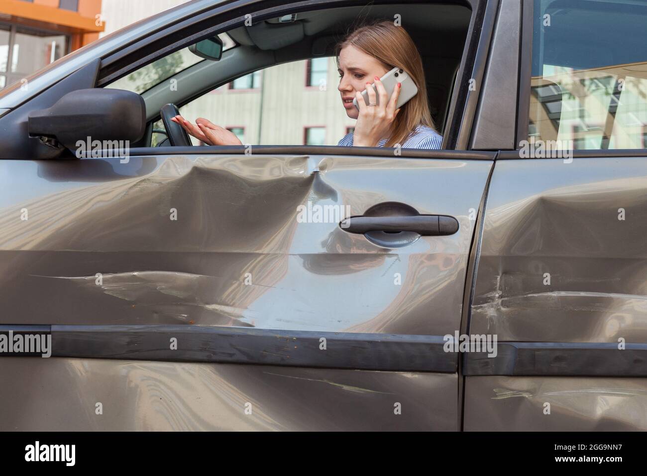 Side view of upset woman with blonde hair sitting in crashed auto and talking phone, calling to insurance agent or to husband, has problems with car d Stock Photo