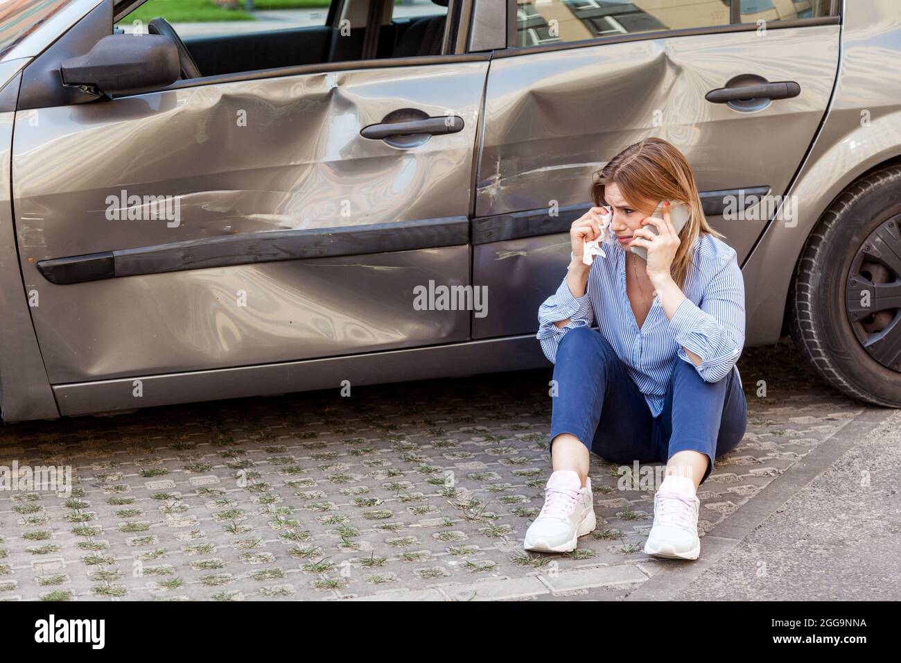 Portrait of blonde female crying and talking phone, crashed side part of her automobile and being in despair, calling asking for help or insurance ins Stock Photo