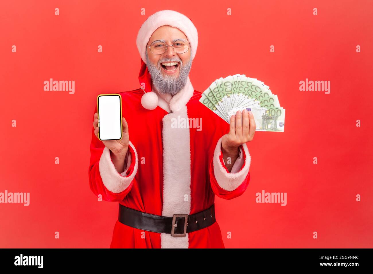 Smiling satisfied elderly man with gray beard wearing santa claus costume showing euro banknotes and smartphone with blank white screen. Indoor studio Stock Photo