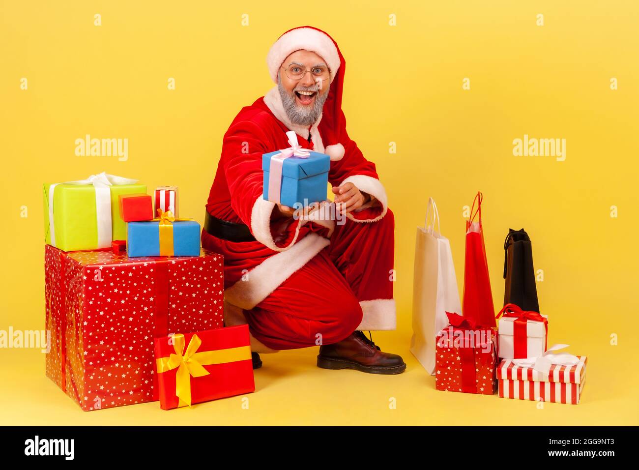 Smiling amazed elderly man with gray beard in santa claus costume sitting near wrapped present boxes, and giving gift, congratulating with New year. I Stock Photo