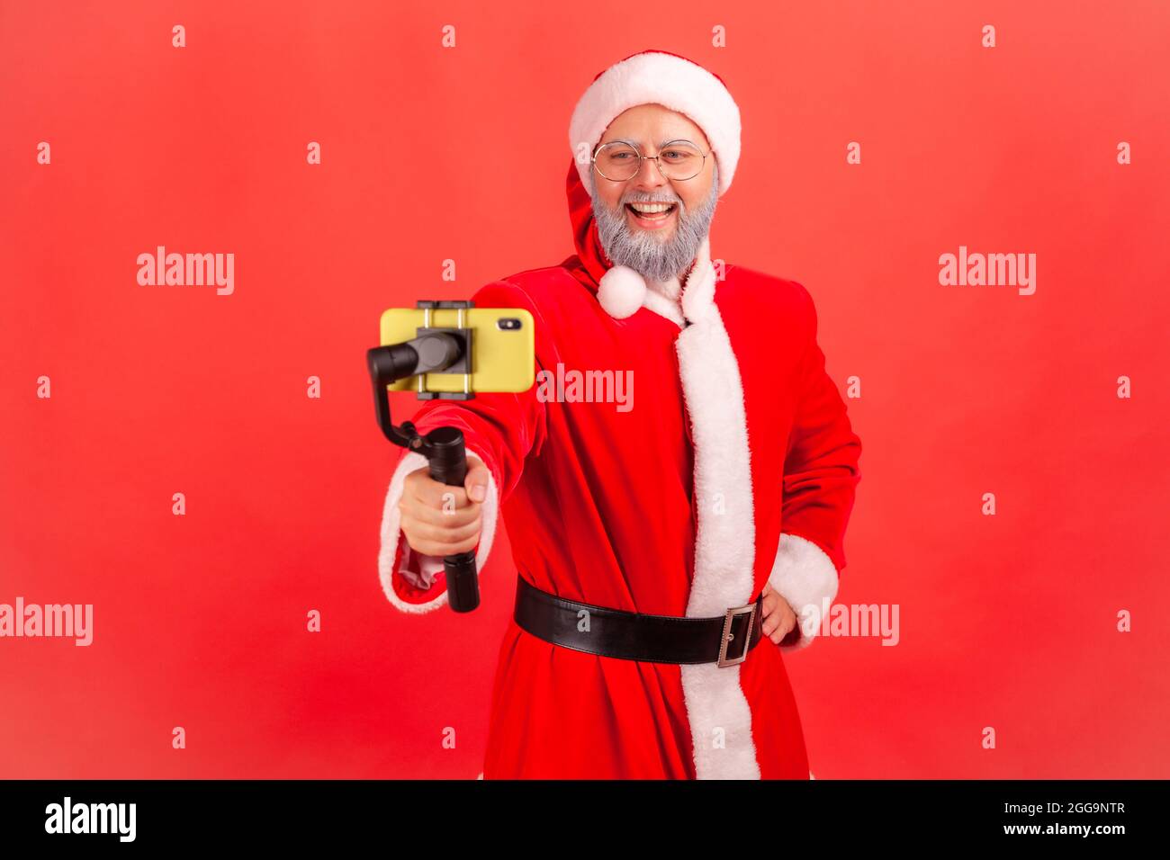 Attractive elderly man with gray beard wearing santa claus costume holding steadicam, broadcasting livestream, blogger talking with followers. Indoor Stock Photo