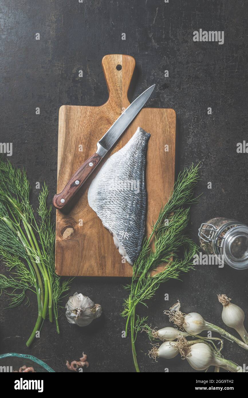 Dorado fillet on cutting board with  kitchen knife and bunch of dill on dark kitchen table with other ingredients for delicious fish cooking. View fro Stock Photo