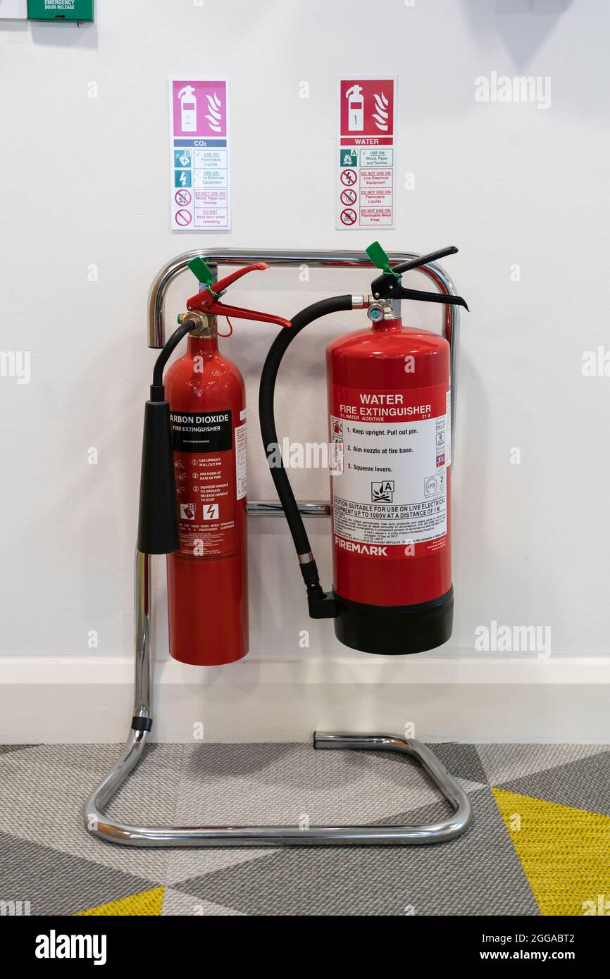 A pair of red fire extinguishers - water and carbon dioxide - on a support rack in an office with safety notices / signs, UK Stock Photo