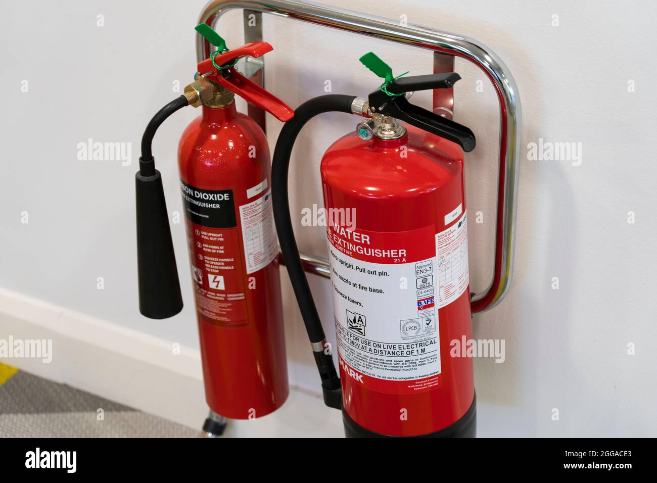 A pair of red fire extinguishers - water and carbon dioxide - on a support rack in an office, UK Stock Photo