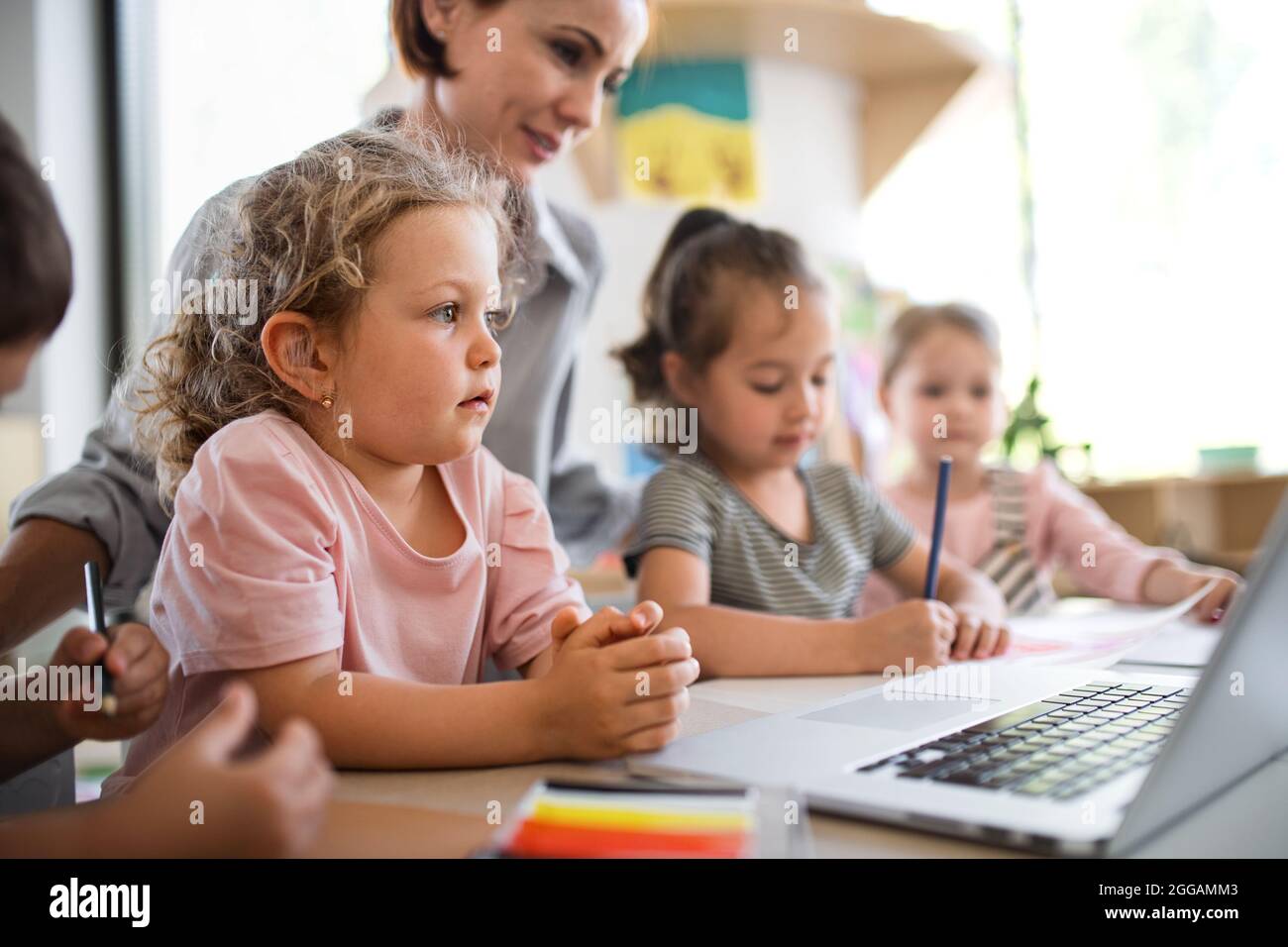 Group of small nursery school children with teacher indoors in classroom, using laptop. Stock Photo