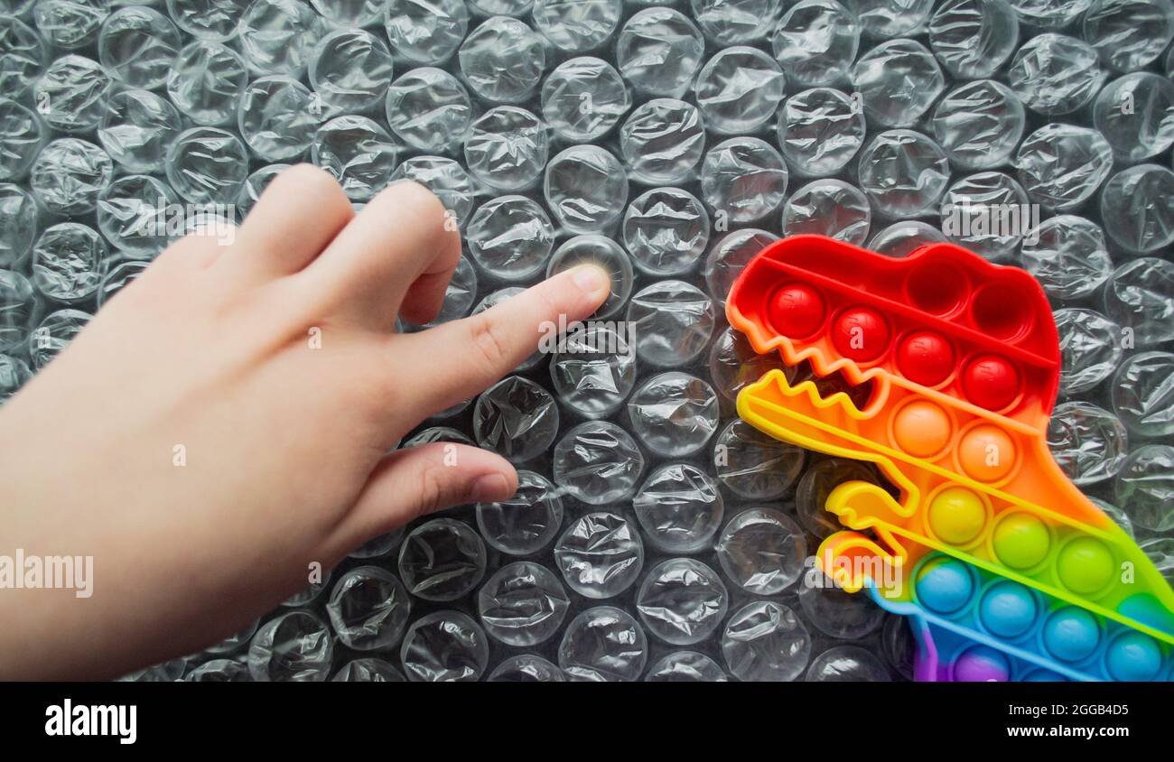 Toy pop it dinosaur rainbow colors on a bubble wrap background. Kid finger pushes bubble, dino looks at it. Copy space. Stock Photo